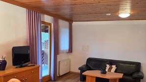 Un salon meublé avec un plancher en bois, un canapé en cuir noir, une table basse en bois, un téléviseur posé sur un meuble en bois et une fenêtre avec des rideaux.
