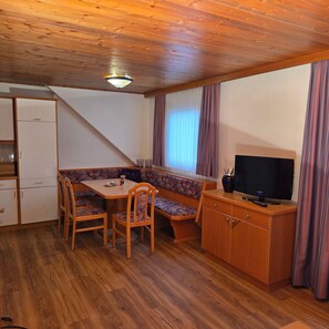 Una stanza compatta con soffitto in legno, un tavolo da pranzo con sedie, una panca, un televisore su un mobile in legno e una finestra con tende.