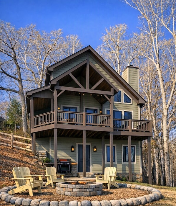 A two-story house with a balcony, a stone fire pit, and Adirondack chairs. - Peacful 3-bedroom cabin in Pigeon Forge with Mountain View, Hot Tub, Fire Pit (Sevierville)