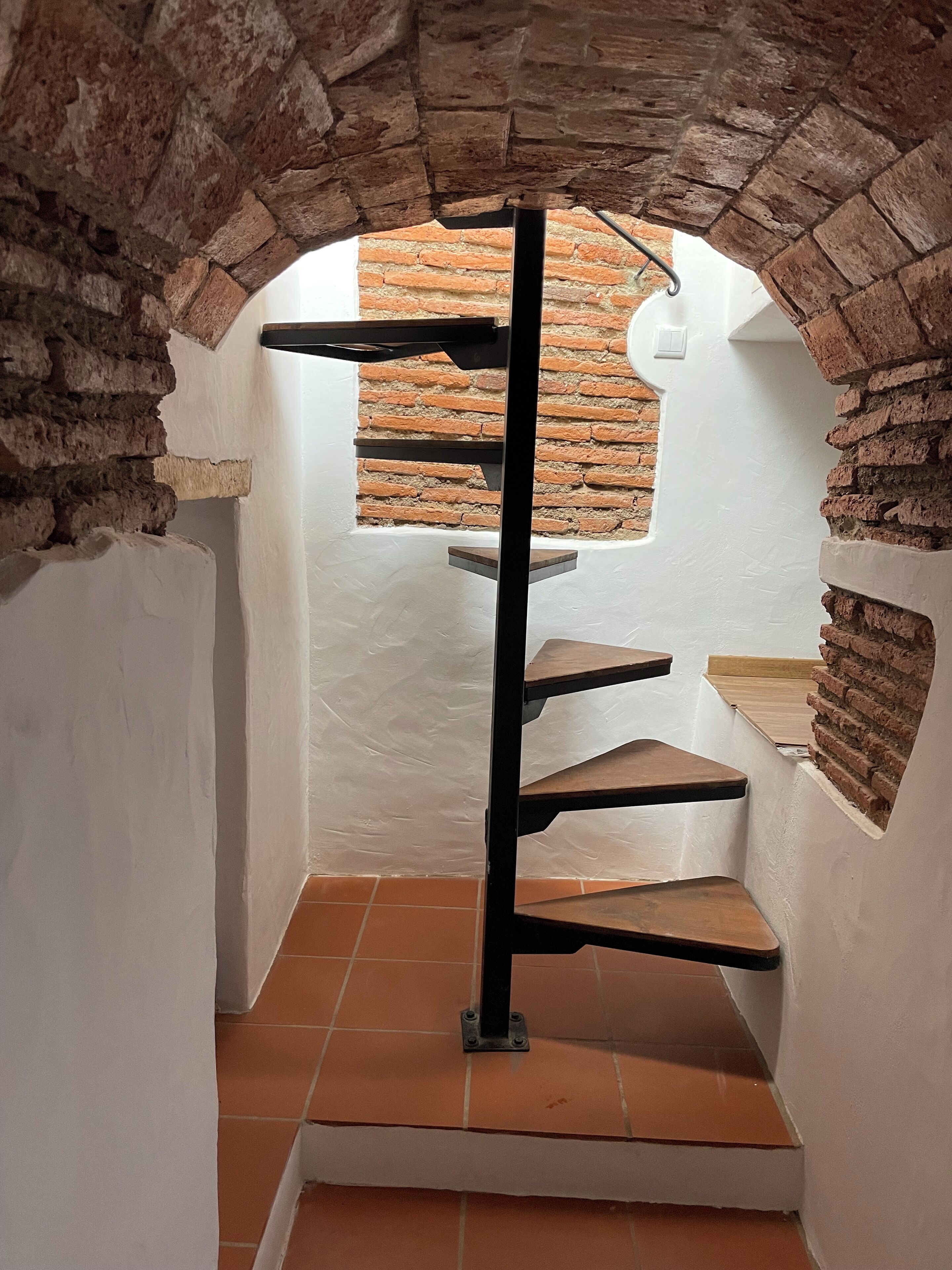 A narrow staircase with a black metal railing, brick walls, and a small window with a brick pattern.