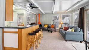 Et moderne køkken med en bar, et siddeområde med sofa og stole, samt store vinduer med persienner.