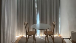 Ein Zimmer mit einem runden Tisch, zwei Stühlen, einem Fenster mit transparenten Vorhängen und einer Lampe auf einem Nachttisch.