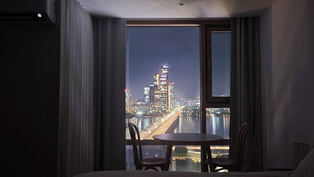 Ein Hotelzimmer mit großem Fenster, das einen nächtlichen Blick auf die Stadt bietet, einem Bett und einem kleinen Tisch mit Stühlen.