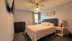 Une chambre à coucher comprenant un lit, une table de chevet, une lampe, une commode, un ventilateur de plafond et un téléviseur fixé au mur.