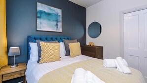 A neatly made bed with a patterned bedspread, a blue upholstered headboard, and a bedside table with a lamp. - Charming City Centre 3 Bed Flat, High St Location! (Chester)