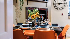 A dining area with a table set for four, featuring orange chairs, a large wall clock, and a vase with sunflowers. - Charming City Centre 3 Bed Flat, High St Location! (Chester)