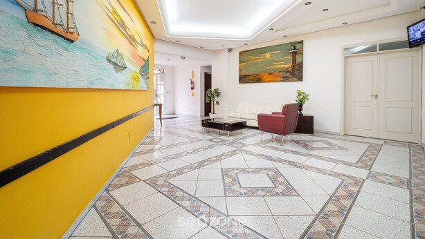 A hotel lobby with a tiled floor, a reception desk, a red sofa, a TV, and a painting of a ship on the wall. - Apartments 230m From the Beach in Ingleses - ZIO (Florianópolis)