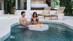 Un área de piscina interior moderna con dos sillones de descanso, una mesita con fruta y una cama al fondo.