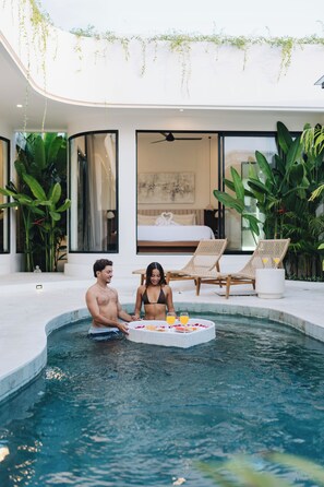 Ein moderner Poolbereich im Innenbereich mit zwei Liegestühlen, einem kleinen Tisch mit Obst und einem Bett im Hintergrund.