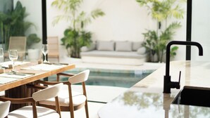 Un comedor moderno con una mesa de madera dispuesta para dos personas, un grifo negro, y vistas a una piscina y follaje al fondo.