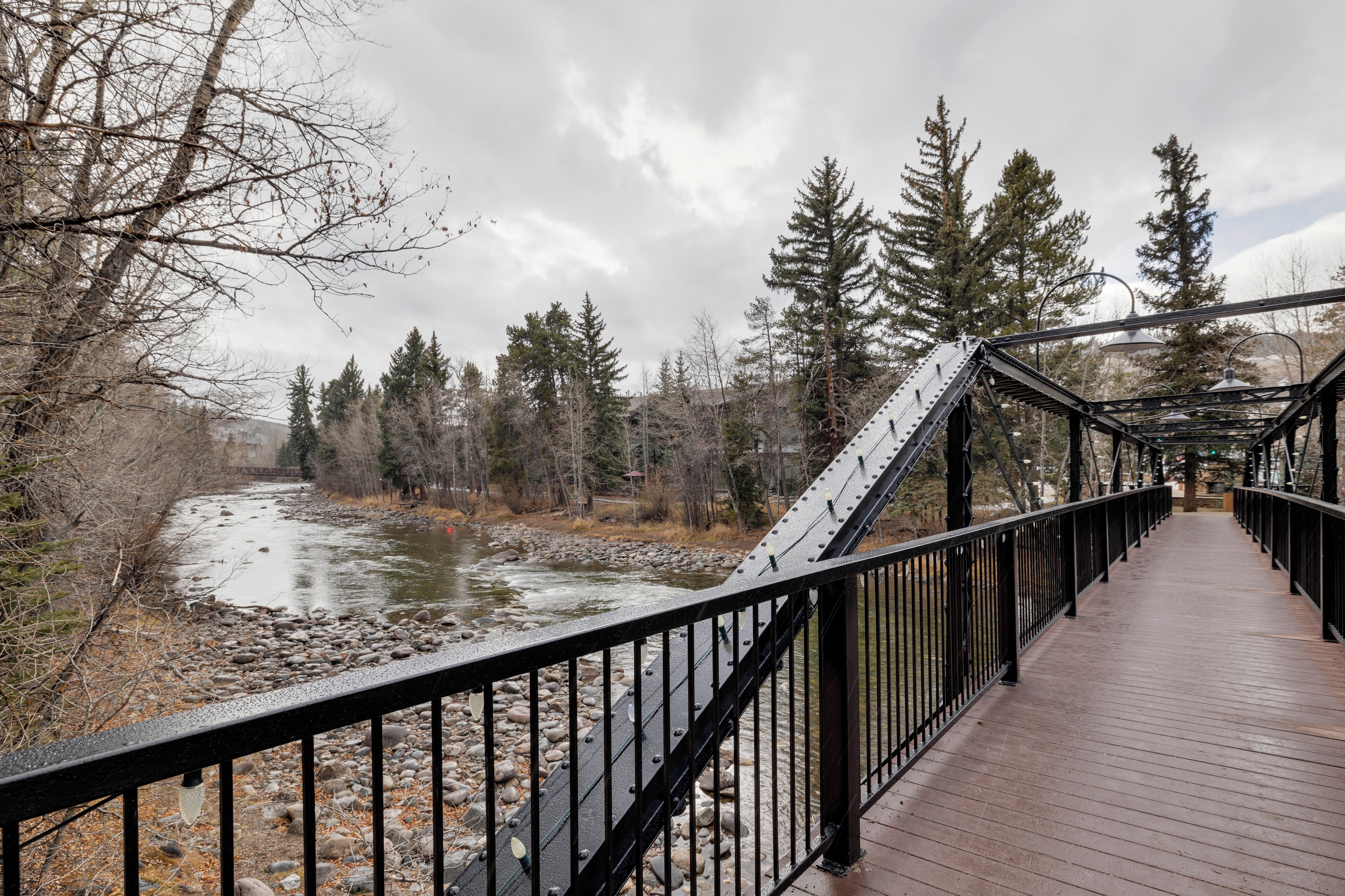A wooden bridge with metal railings over a river, surrounded by trees and a cloudy sky.
