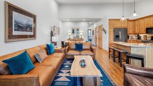 A spacious living room with a brown leather sofa, a wooden coffee table, and a kitchen area with wooden cabinets and a stainless steel refrigerator.