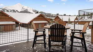 En träveranda med stolar och ett bord, med utsikt över ett snötäckt berg och flera stugor.