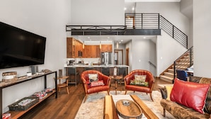 Amplia sala de estar con techo alto, una cocina al fondo y una escalera que conduce a un nivel superior.