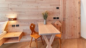 Une pièce aux murs lambrissés, contenant un lit, une table, deux chaises, un vase avec des plantes séchées et une porte.
