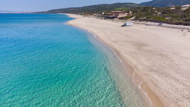 A coastal beach with clear turquoise water, sandy shore, and buildings in the background. - Comfy Sardinian Retreat With Pool Access (Viddalba)