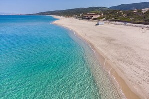 Ein Küstenstrand mit klarem türkisblauem Wasser, sandigem Ufer und Gebäuden im Hintergrund.