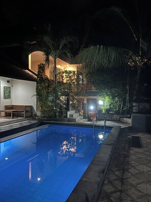 En swimmingpool med fliseterrasse, omgivet af palmer og en bygning med flisebelagt tag.