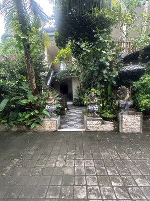 A courtyard with a tiled pathway leading to a building entrance, surrounded by lush greenery and stone statues.