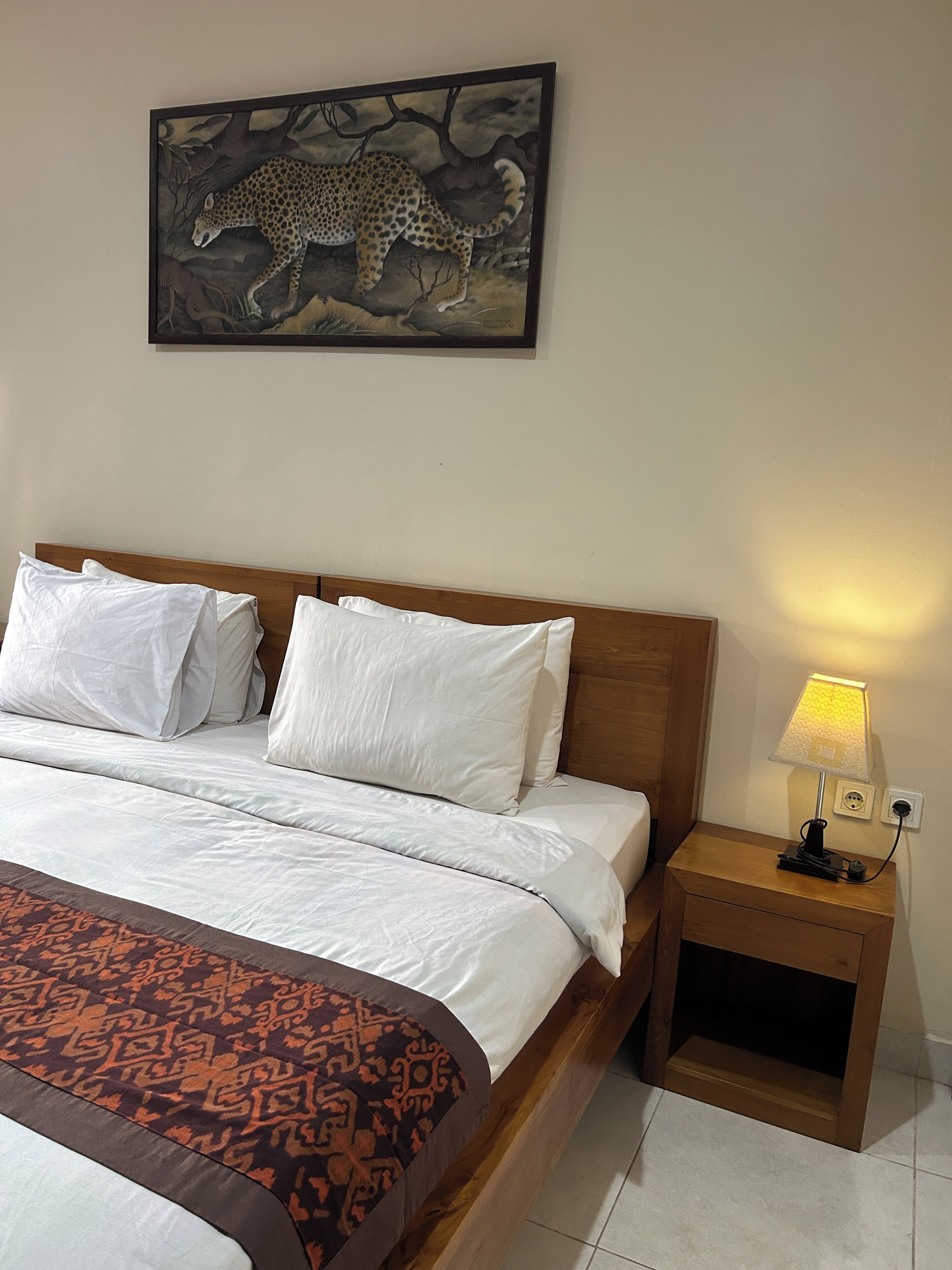 A neatly made bed with white bedding, a patterned throw, a nightstand with a lamp, and a leopard painting on the wall.