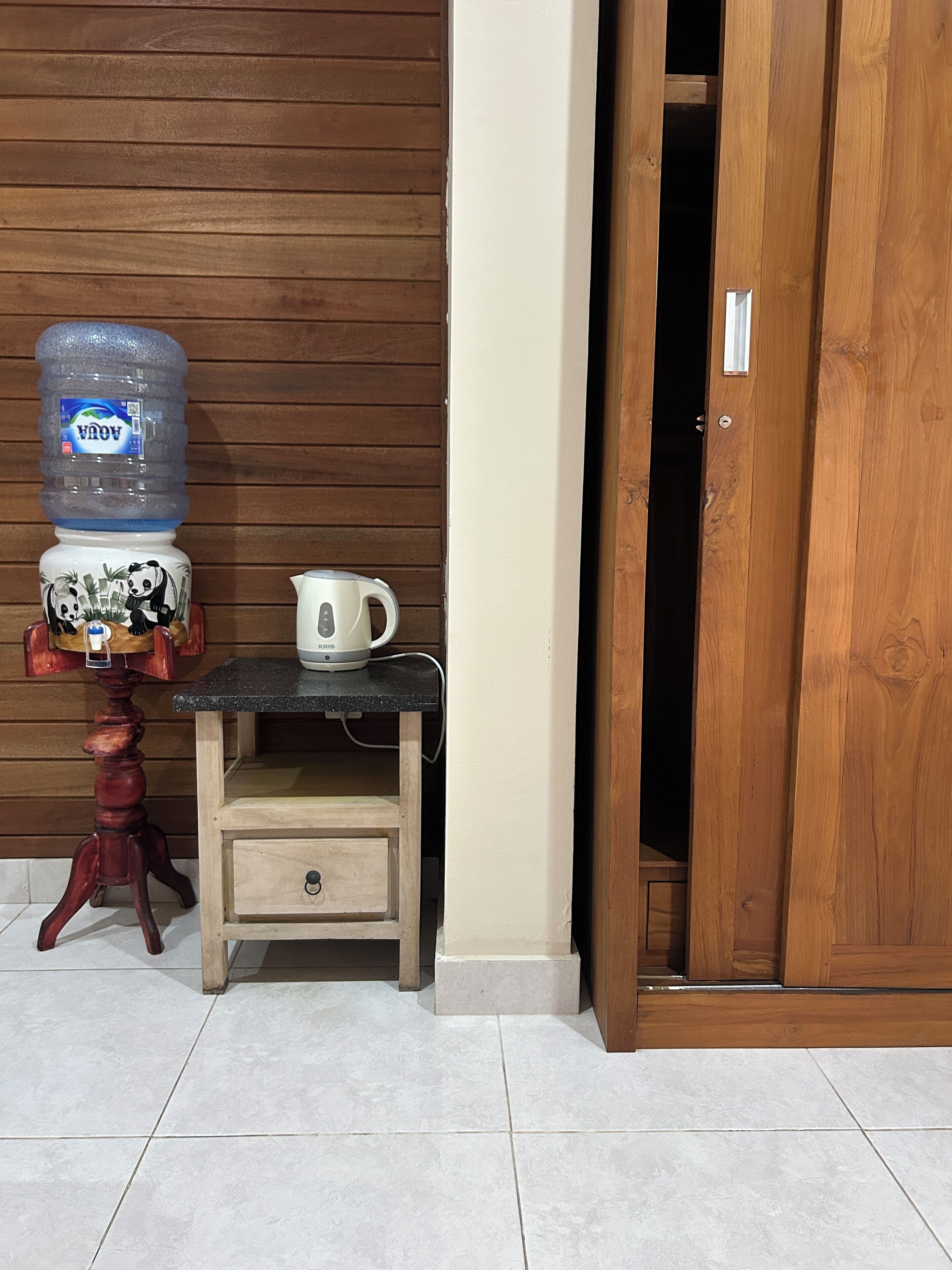 Una pequeña mesa de madera con un dispensador de agua y una tetera, un armario de madera con un cajón, y una puerta de madera.