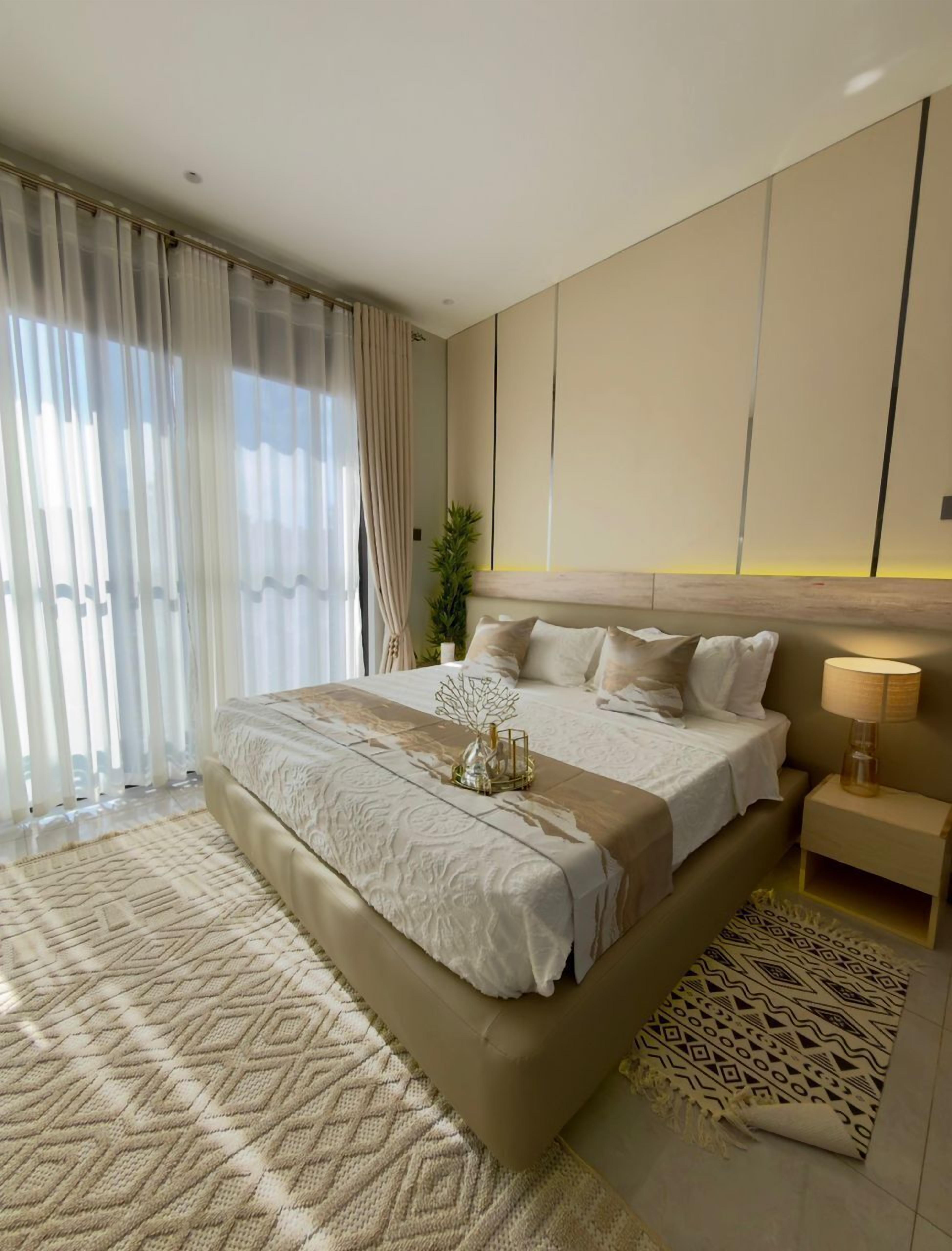 A modern bedroom with a large bed, a bedside table with a lamp, and a view of the outdoors through sheer curtains.