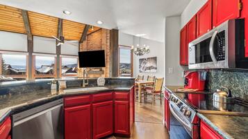 A modern kitchen with red cabinetry, a large island, stainless steel appliances, and a view of the mountains through large windows.