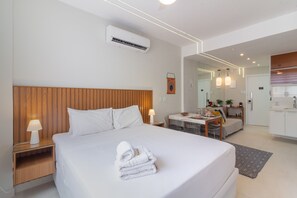 A modern hotel room with a wooden headboard, a neatly made bed with white linens, a small bedside table with a lamp, a dining area with a table and chairs, and a kitchenette in the background. - 5 Minutes From Copacabana Beach B703 (Rio de Janeiro)