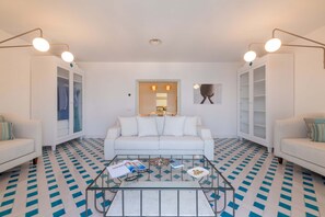 Ein modernes Wohnzimmer mit einem weißen Sofa, einem Glastisch und einem gemusterten Boden. Zwei weiße Sessel und ein weißes Schrank mit Glastüren ergänzen das Bild.
