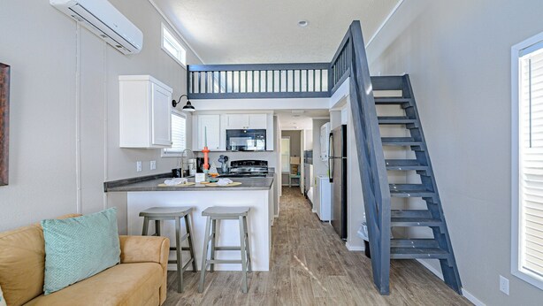 A modern kitchen with a central island, a dining area with two bar stools, a staircase, and a living room with a sofa and a decorative pillow.