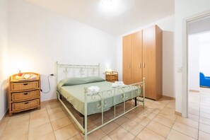 A bedroom with a metal bed frame, a green bedspread, a wooden wardrobe, wicker furniture, and a tiled floor. - Relaxing Apartment Close to Santa Lucia Beach (Siniscola)