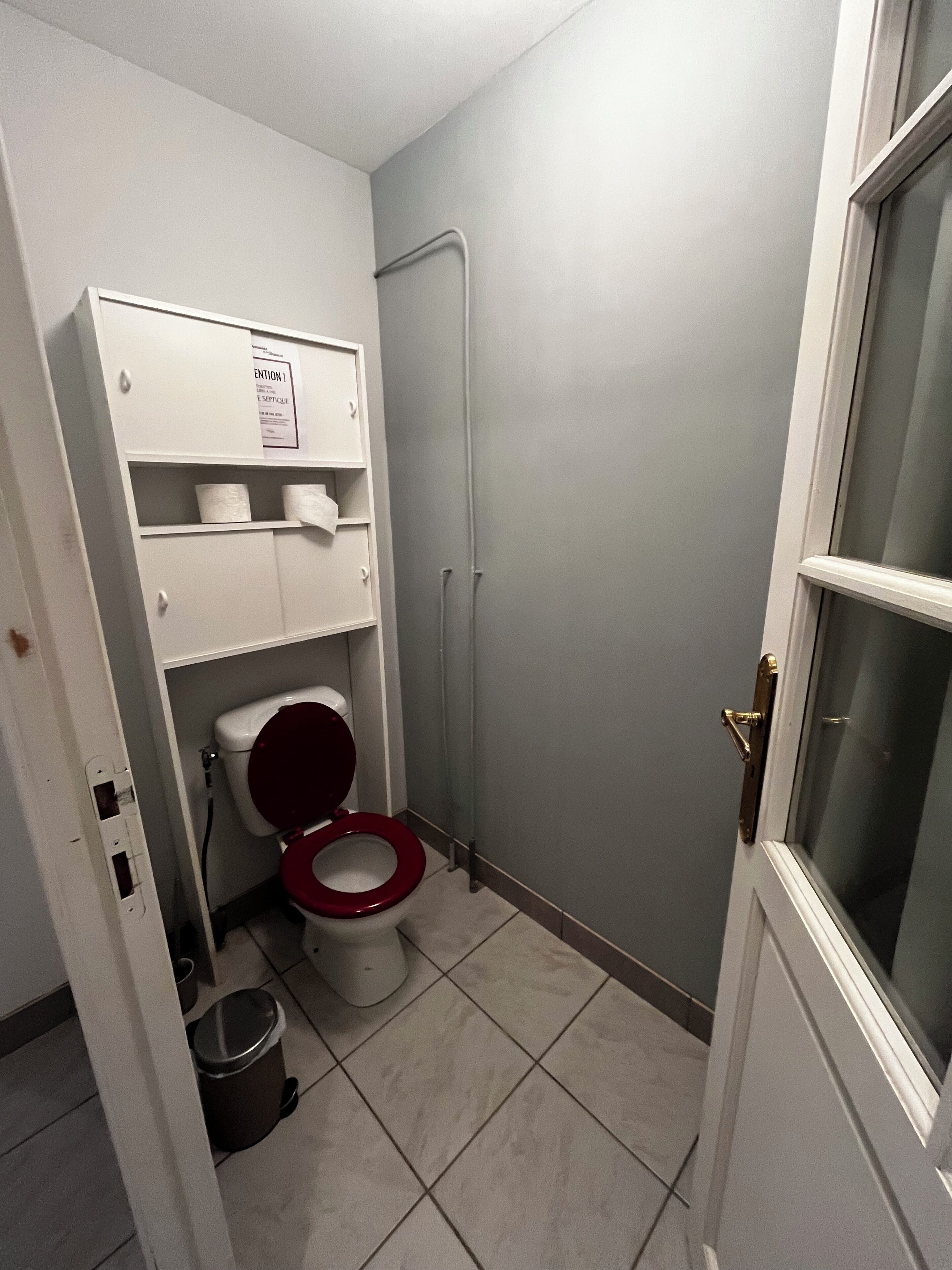 Un baño pequeño con inodoro, estante con papel higiénico, bote de basura y zona de ducha.
