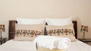 Una cama bien hecha con almohadas blancas y estampadas, una toalla enrollada en una cesta y dos lámparas de noche sobre mesitas de madera.