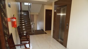 A hotel lobby with a marble floor, wooden chairs, a staircase, and an elevator. - Hotel Nilax (Jaipur)