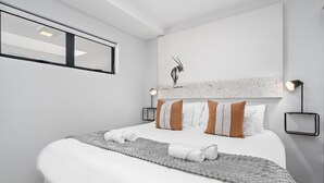 A modern bedroom with a large bed, white bedding, a grey throw blanket, and a wall-mounted lamp. - Beach Break 17 (Cape Town)