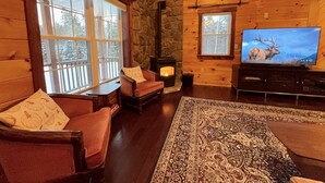 L’intérieur chaleureux d’un chalet avec une cheminée en pierre, des murs en bois, une grande fenêtre et un téléviseur diffusant une image d’un élan.