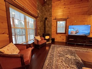 A cozy cabin interior with a stone fireplace, wooden walls, a large window, and a television displaying an elk. - Private, Comfy Cabin in Rangeley/Trails, Lakes, Fire Pit/Sleeps 8/Starry Sky (Rangeley)
