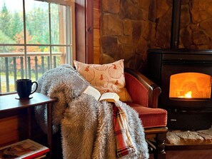 A cozy room with a fireplace, a window with a view of trees, and a plush chair with a blanket.