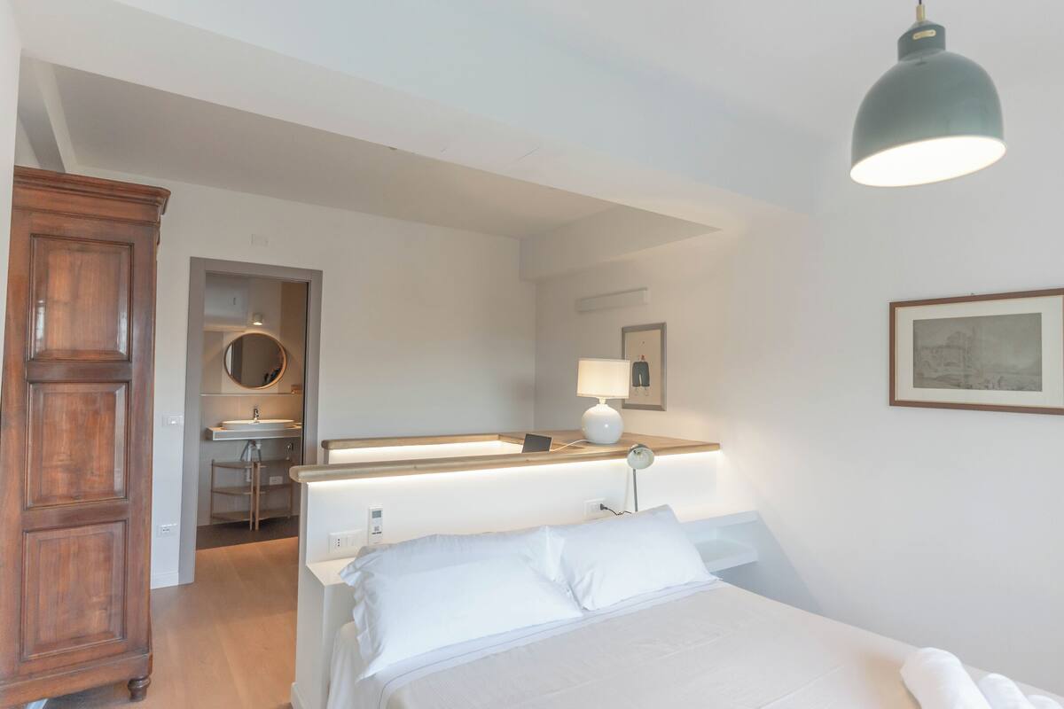 A bedroom with a bed, a wooden wardrobe, a bathroom with a sink, mirror, and a round mirror.