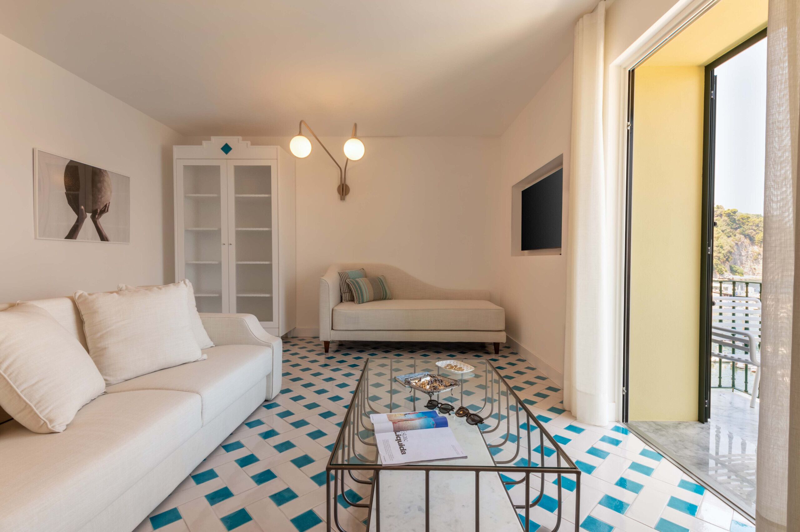 A modern living room with a white sofa, a glass coffee table with a magazine and sunglasses, a white cabinet, and a patterned floor.