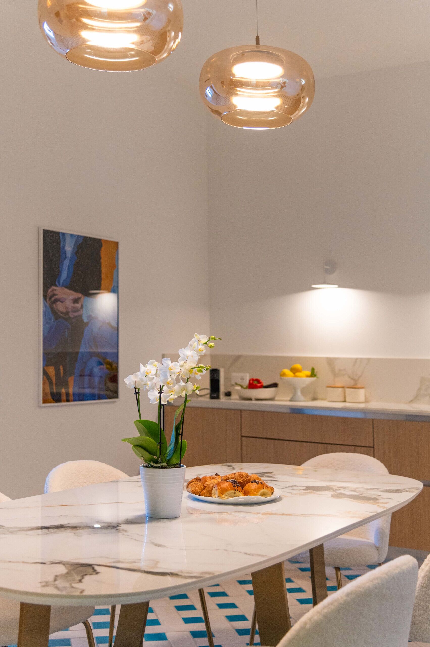 Uma sala de jantar moderna com uma mesa de mármore, cadeiras brancas, uma orquídea em vaso e um prato de croissants.