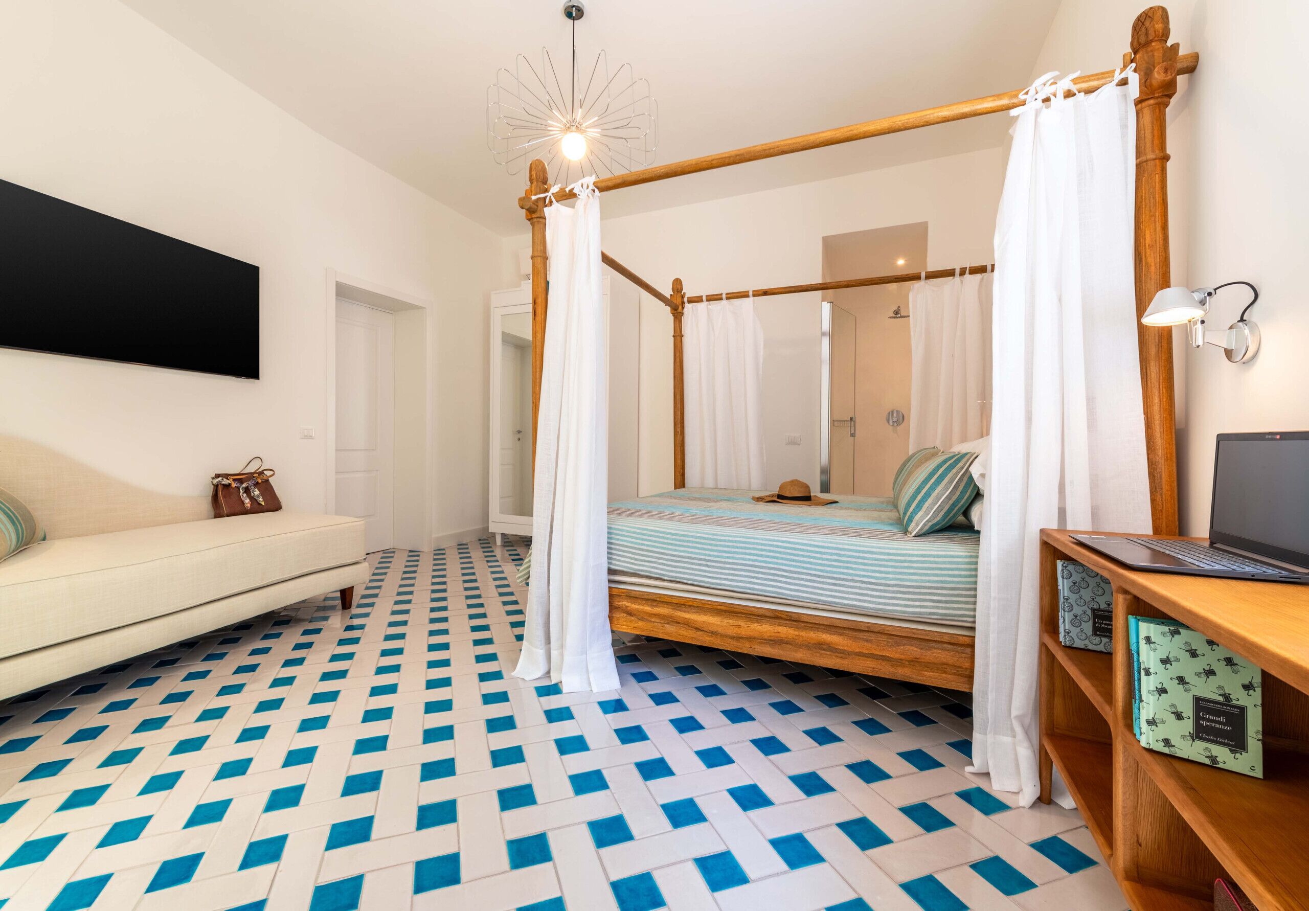 A bedroom with a four-poster bed, a wooden desk with a laptop, a bookshelf, and a patterned floor.