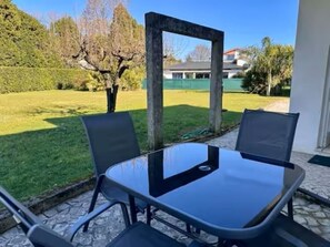 Mesa de vidrio con cuatro sillas, ubicada en un patio de piedra con vistas a un área de césped y árboles.