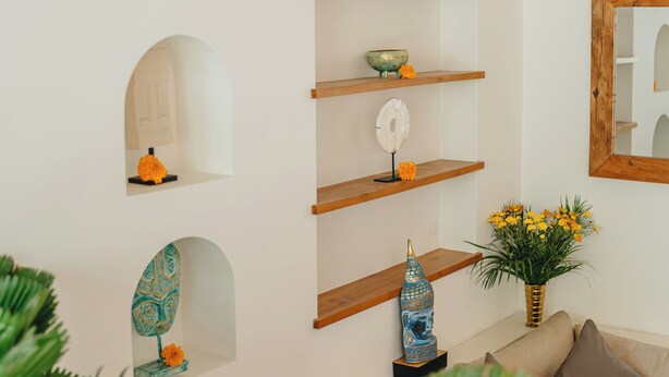 En stue med hvid væg, træhylder, en Buddha-statue, et spejl og en vase med blomster.