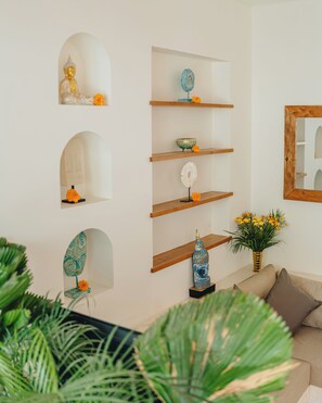 Ein Wohnzimmer mit weißer Wand, Holzregalen, einer Buddha-Statue, einem Spiegel und einer Vase mit Blumen.