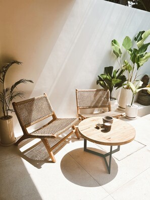 Eine sonnendurchflutete Terrasse mit zwei Korbstühlen und einem runden Holztisch, umgeben von Topfpflanzen und einer Buddha-Statue.