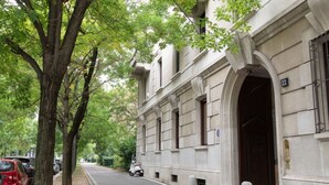 Vista de la calle con un edificio que tiene una gran entrada en arco, coches aparcados y árboles a lo largo de la acera.