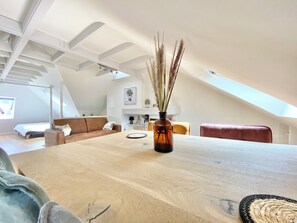 Een ruime kamer met een grote houten tafel, een vaas met gedroogde planten en een hoog plafond met zichtbare balken.