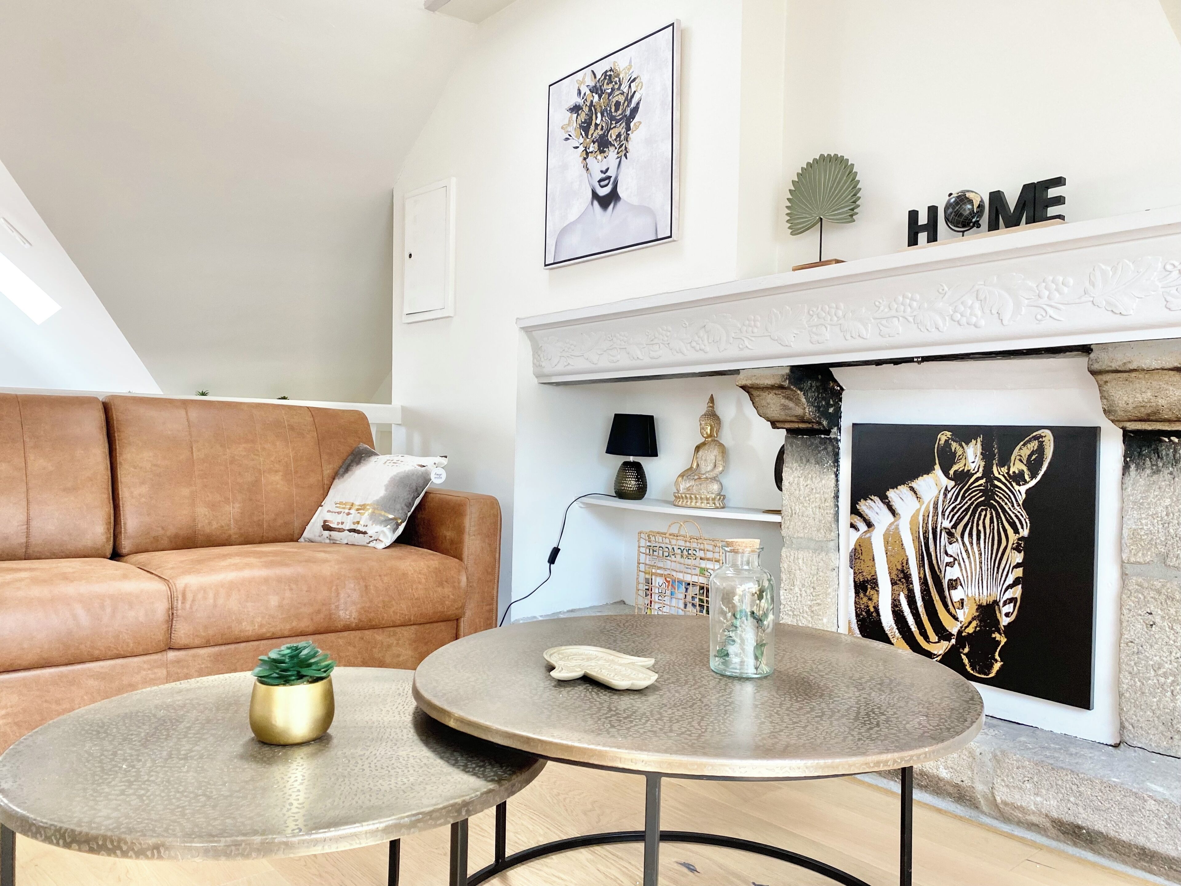 A modern living room with a brown leather sofa, two round tables, a fireplace, and various decorative items including a zebra painting and a Buddha statue.