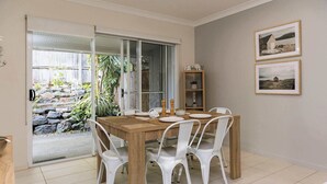 A dining area with a wooden table and white chairs, a sliding glass door leading to a garden, and two framed pictures on the wall.